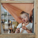 Child standing behind the wooden window frame playing with the grey bear puppet and the orange fox puppet. 