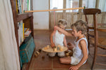 2 children sitting on a terracotta floor playing with the Grapat wooden pendulum set balancing on a wooden chair and bookshelf with wooden blocks on the wooden balance board and scattered on the terracotta floor