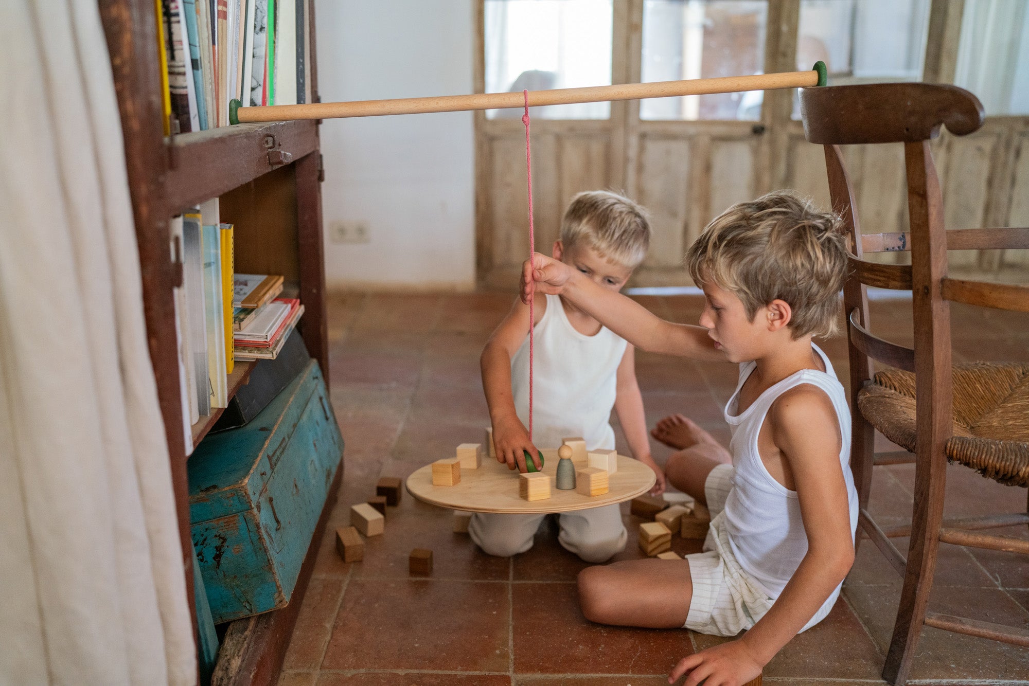 2 children sitting on a terracotta floor playing with the Grapat wooden pendulum set balancing on a wooden chair and bookshelf with wooden blocks on the wooden balance board and scattered on the terracotta floor