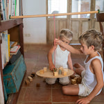 2 children sitting on a terracotta floor playing with the Grapat wooden pendulum set balancing on a wooden chair and bookshelf with wooden blocks on the wooden balance board and scattered on the terracotta floor