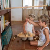 2 children sitting on a terracotta floor playing with the Grapat wooden pendulum set balancing on a wooden chair and bookshelf with wooden blocks on the wooden balance board and scattered on the terracotta floor