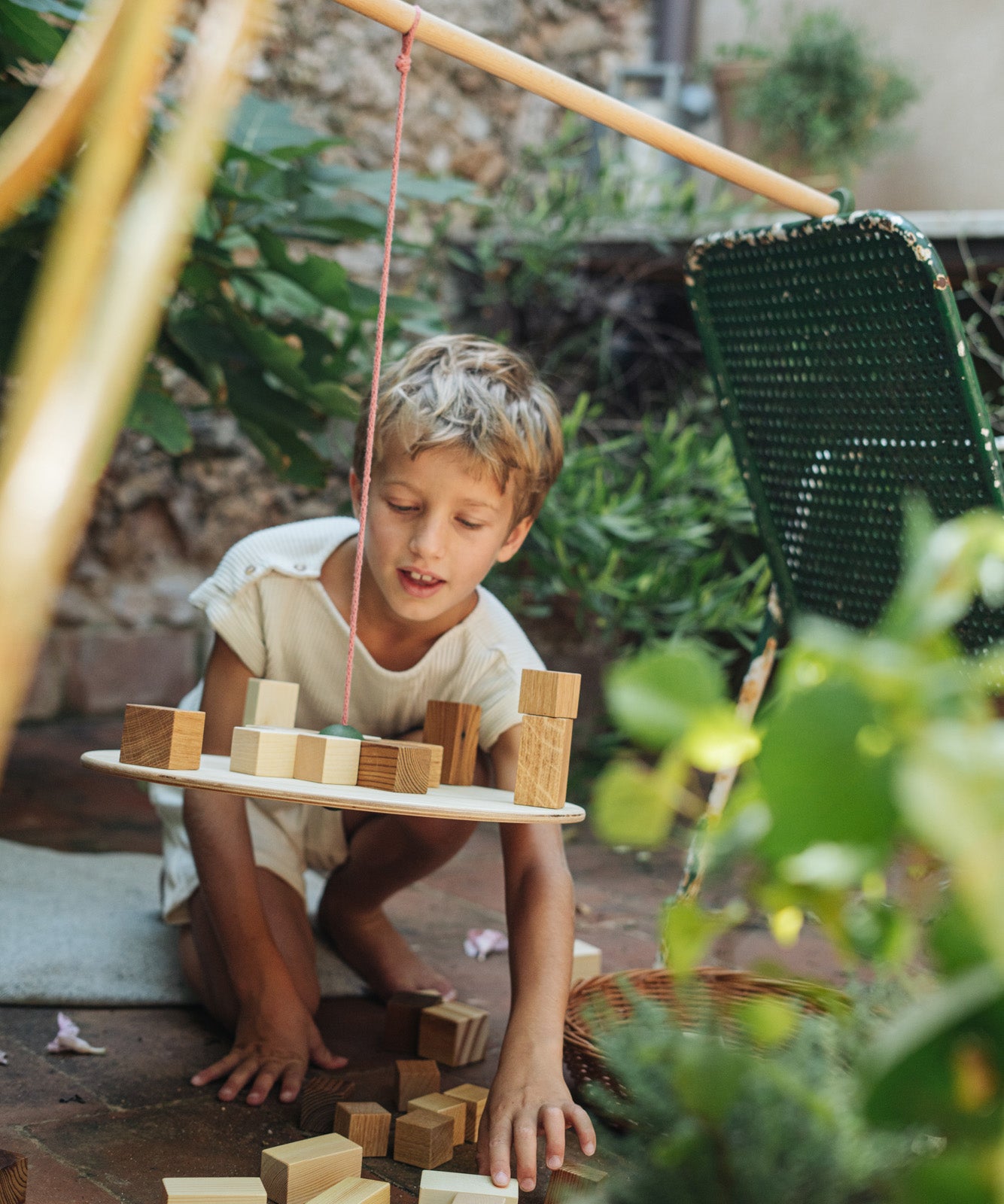 Child playing in the garden with the Grapat wooden pendulum set resting on a garden chair and wooden clothes horse. Showing the handcrafted wooden blocks balancing on the balance board and scattered on the stone floor.