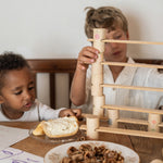 2 children sitting next to a wooden table stacking the handcrafted Grapat wooden tubes and planks. A close up showing the different painted tubes featuring flowers, windows and ladders. 