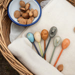 Grapat handcrafted wooden twin soul puppets on a cream blanket in a woven basket next to a bowl of nuts. Showing the 6 different handcrafted coloured wooden puppets.