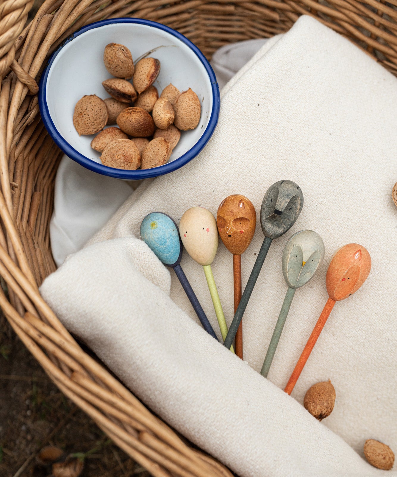 Grapat handcrafted wooden twin soul puppets on a cream blanket in a woven basket next to a bowl of nuts. Showing the 6 different handcrafted coloured wooden puppets.