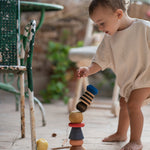 Child playing with the Grapat wooden serendipity stacking set outside on the floor next to a garden chair . Showing the different handcrafted tactile pieces stacked on the stone floor. Features a brown, light pink, blue yellow orange and striped pieces. 