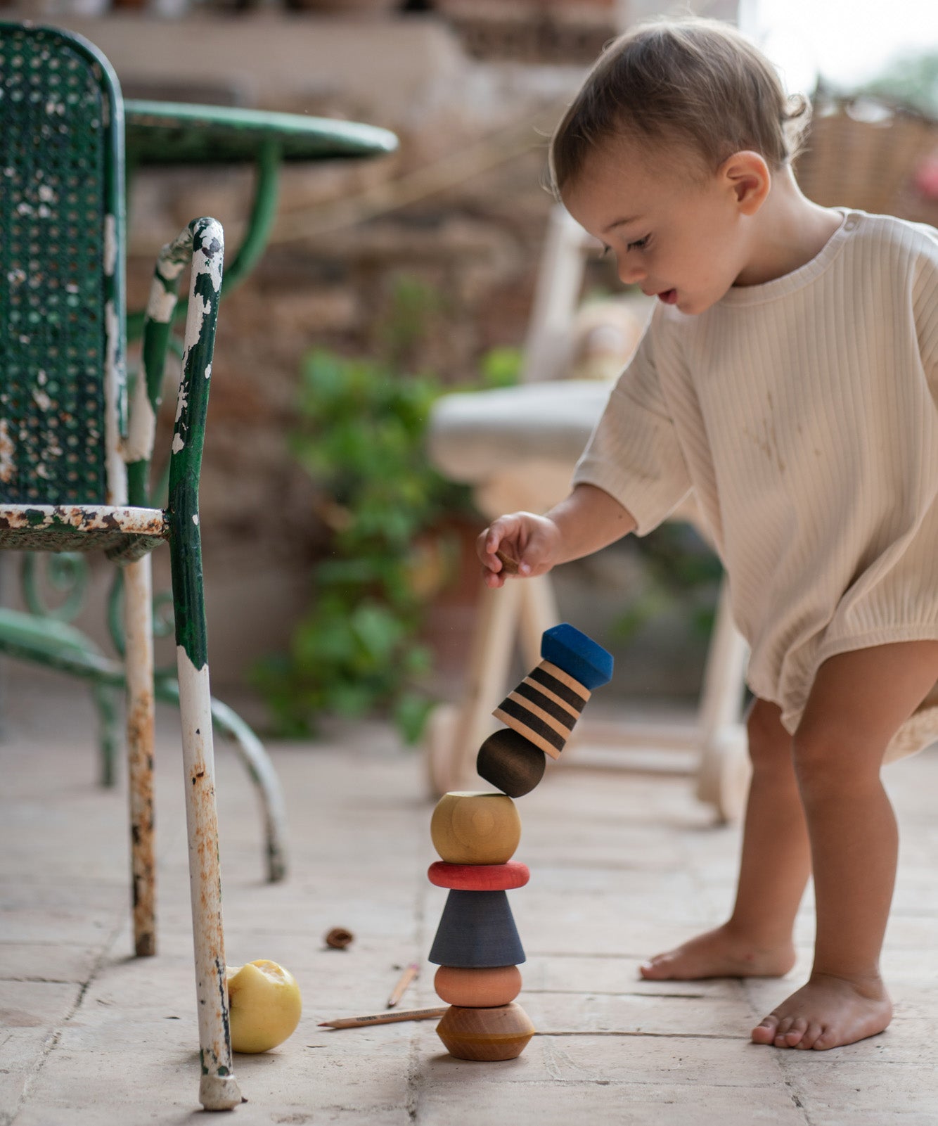 Child playing with the Grapat wooden serendipity stacking set outside on the floor next to a garden chair . Showing the different handcrafted tactile pieces stacked on the stone floor. Features a brown, light pink, blue yellow orange and striped pieces. 