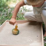 A child in the garden playing with the Grapat wooden yellow flowing bird in a Grapat freeplay tray filled with sand on a gravel floor