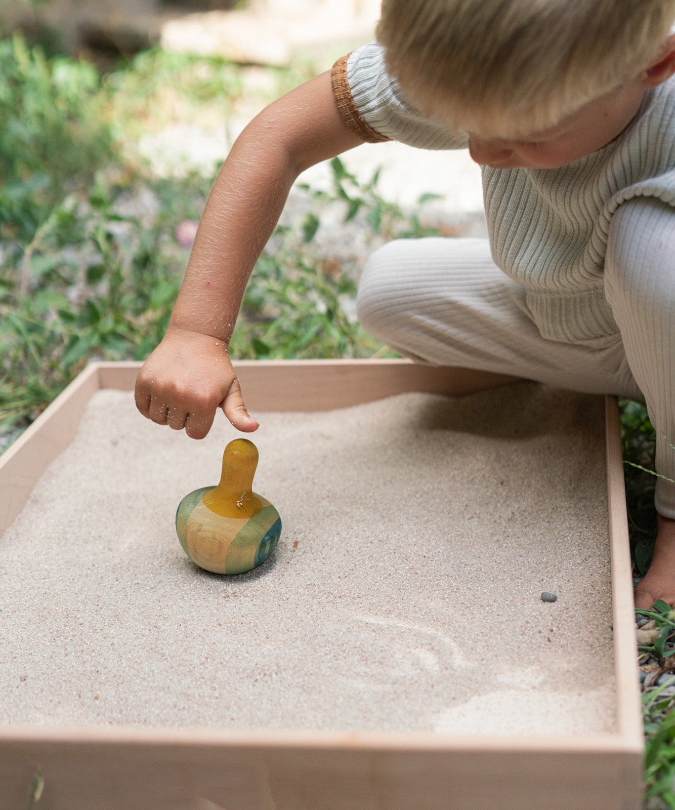 A child in the garden playing with the Grapat wooden yellow flowing bird in a Grapat freeplay tray filled with sand on a gravel floor