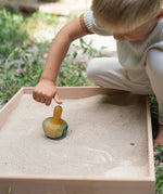 A child in the garden playing with the Grapat wooden yellow flowing bird in a Grapat freeplay tray filled with sand on a gravel floor
