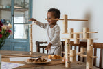Child standing on a wooden chair next to a dining table balancing a handcrafted wooden plank on a tower of woode tubes from the Grapat wooden woodland set.