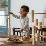 Child standing on a wooden chair next to a dining table balancing a handcrafted wooden plank on a tower of woode tubes from the Grapat wooden woodland set.
