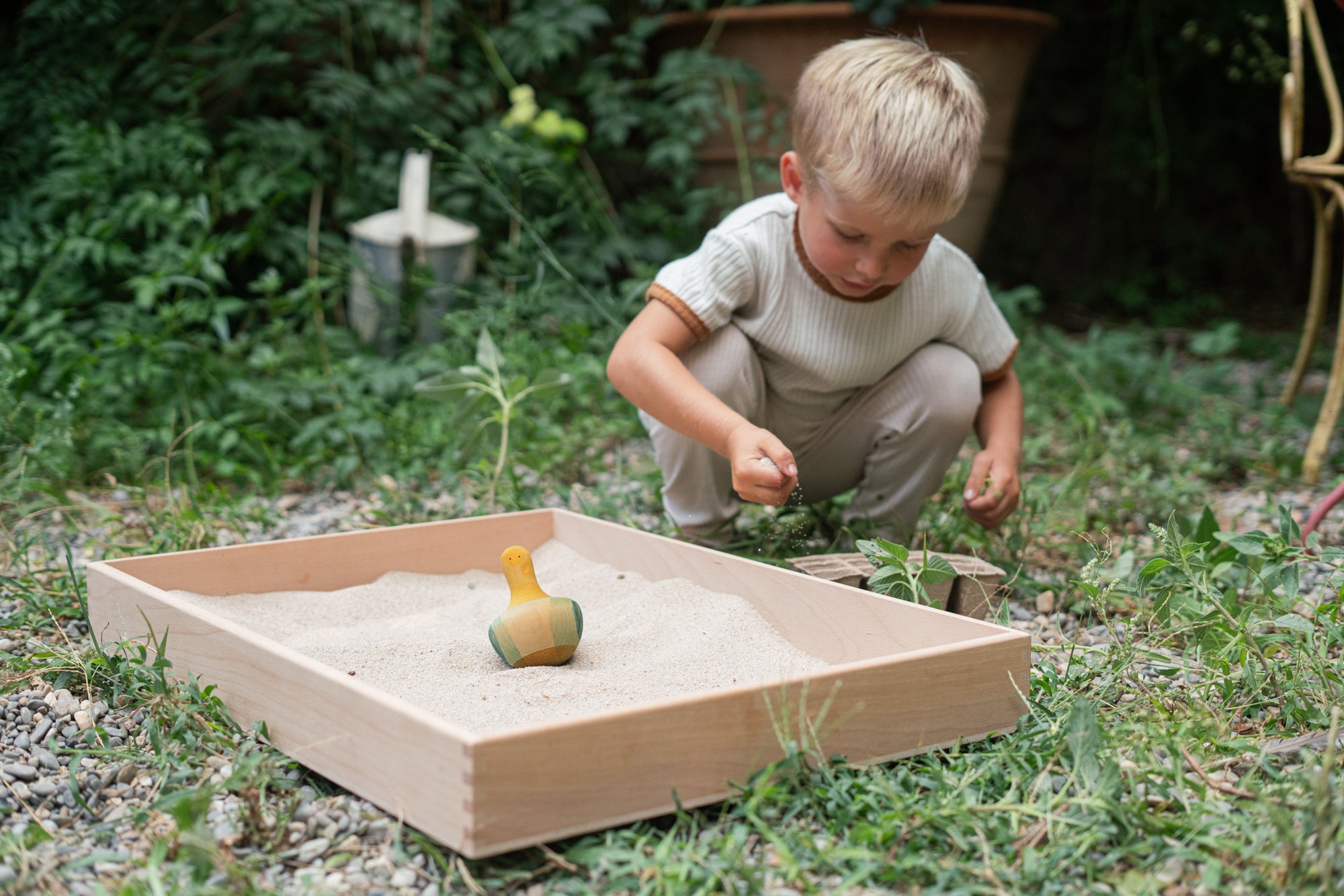 Child playing in the garden with the Grapat wooden flowing yellow bird sitting in the Grapat wooden freeplay tray filled with sand on a grassy floor. 