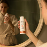 An adult standing in front of a mirror and holding the Green People glow & protect spf30 150ml cream bottle