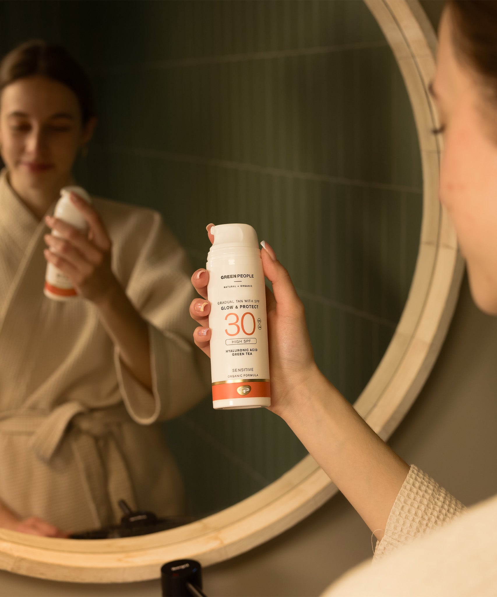 An adult standing in front of a mirror and holding the Green People glow & protect spf30 150ml cream bottle
