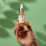 A person holding the Green People Age Defy+ Liquid Gold Tanning Drops 30ml bottle in their hand on a green background