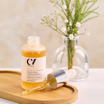 Green People Age Defy+ Liquid Gold Tanning Drops 30ml stood on a wooden tray with a plant in the background, showing the dropper