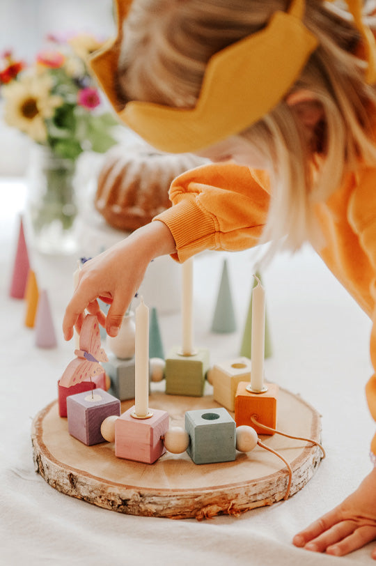 Child wearing a yellow crown and jumper playing with a Grimm's pastel wooden celebration ring with beeswax candles and lilac butterfly decorative figure to represent Grimm's family celebrations at Babipur, official UK stockist.