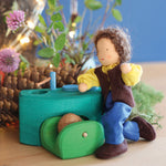 Grimm's Brown Haired Man Doll - Erle is bent down on its knees, next to a little green wooden kitchen. There are flowers and thin tree branches in the background.