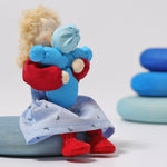 The Grimm's Blonde Woman Doll, is holding a small doll representing a child which is dressed in blue, and is sitting on a blue Grimm's Wooden river pebbles piece