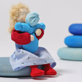 The Grimm's Blonde Woman Doll, is holding a small doll representing a child which is dressed in blue, and is sitting on a blue Grimm's Wooden river pebbles piece