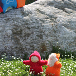 An orange and red Grimm's Dwarf doll enjoying the sunshine and stood on a patch of grass full of small daisies. Behind the dwarf toys is a large grey rock and a Grimm's House