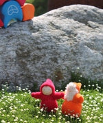 An orange and red Grimm's Dwarf doll enjoying the sunshine and stood on a patch of grass full of small daisies. Behind the dwarf toys is a large grey rock and a Grimm's House