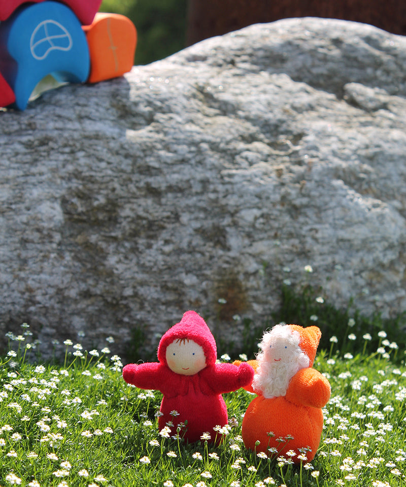 An orange and red Grimm's Dwarf doll enjoying the sunshine and stood on a patch of grass full of small daisies. Behind the dwarf toys is a large grey rock and a Grimm's House