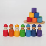 The Grimm's Wooden Friends Peg Dolls all stood in a row, with the Grimm's wooden bowls in the background. The focus is on the peg dolls at the front, with the bowls blurred in the background