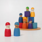 Two Grimm's Rainbow dolls in the foreground with the Wooden Rainbow Bowls and other Grimm's Rainbow Dolls stacked in the background. The focus is on the two peg dolls at the front with the pieces in the background blurred slightly