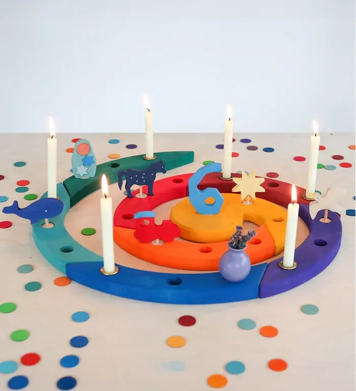 A colorful wooden Grimm's advent spiral celebration ring with built-in candle holders, decorated with small animal and star cutouts and lit white candles, set on a white table covered in colorful confetti.