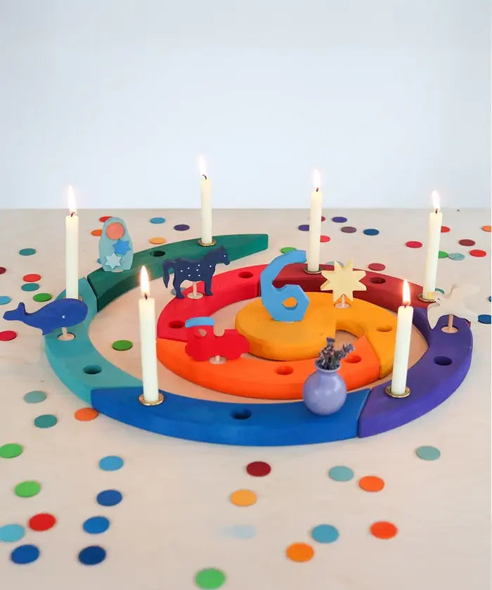 A colorful wooden Grimm's advent spiral celebration ring with built-in candle holders, decorated with small animal and star cutouts and lit white candles, set on a white table covered in colorful confetti.