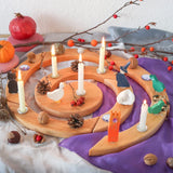 A Grimm's spiral wooden Advent celebration ring decorated with white candles, small ornaments, pinecones, acorns, and colorful bird figurines, styled on a white cloth with a purple fabric backdrop and seasonal props.