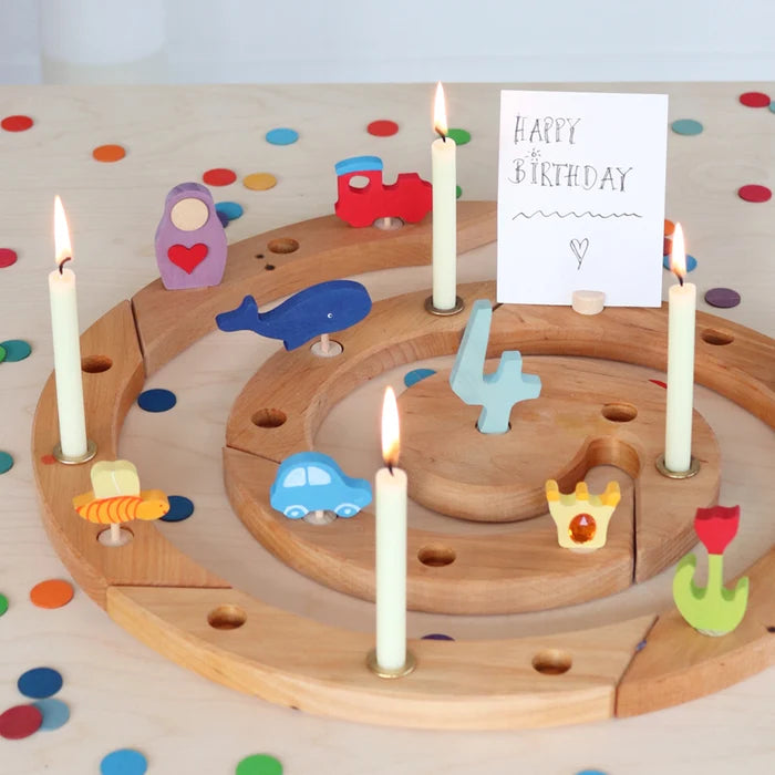 A wooden Grimm's spiral advent celebration ring with colorful toy vehicles and candles, featuring a white card that says 'Happy Birthday' on a decorated tabletop.