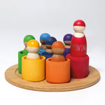 The Grimm's Wooden Rainbow Friends with bowls, stood on a small wooden plate on a cream background. The red bowl has been turned upside down and the red peg doll is stood on top 