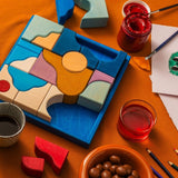 Grimm's wooden wave building set in a blue square frame on a orange cloth next to arts and crafts supplies