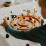 A child's hand interacting with a Grimm's wooden celebration Advent wreath set on a white fabric-covered table. The wooden wreath is decorated with lit taper candles, small wooden angel and star figures, pinecones, acorns, and miniature green trees.