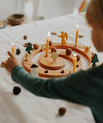 A child's hand interacting with a Grimm's wooden celebration Advent wreath set on a white fabric-covered table. The wooden wreath is decorated with lit taper candles, small wooden angel and star figures, pinecones, acorns, and miniature green trees.