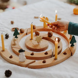 Grimm's natural spiral celebration ring with candles, acorns, pinecones, and small decorative figures arranged on a white fabric surface, with a child's hand holding a lit candle.