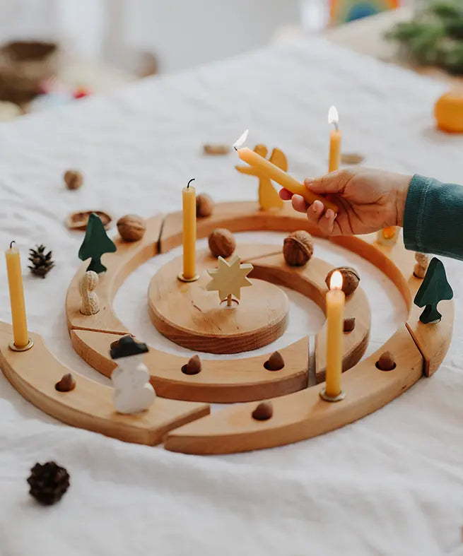 Grimm's natural spiral celebration ring with candles, acorns, pinecones, and small decorative figures arranged on a white fabric surface, with a child's hand holding a lit candle.