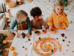 Three children playing with a Grimm's natural spiral celebration ring, acorns, pinecones, and small ornaments in a warm, decorated indoor setting.