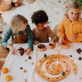 Three children playing with a Grimm's natural spiral celebration ring, acorns, pinecones, and small ornaments in a warm, decorated indoor setting.