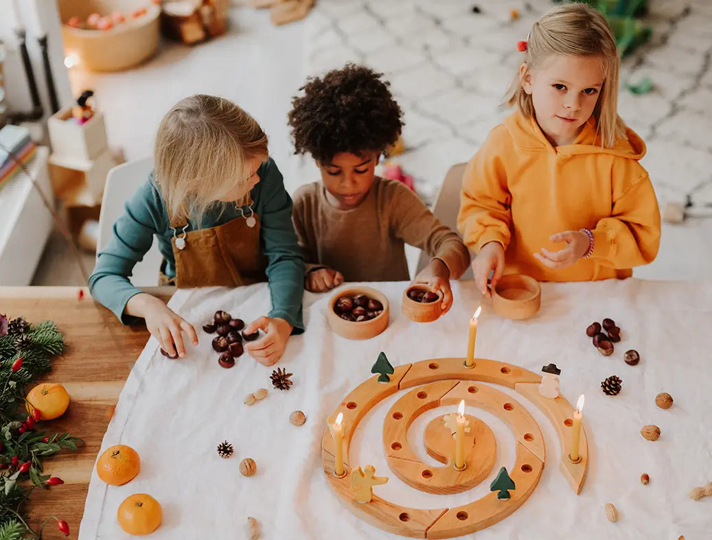 Three children playing with a Grimm's natural spiral celebration ring, acorns, pinecones, and small ornaments in a warm, decorated indoor setting.