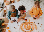Three children sitting around a Grimm's natural spiral celebration ring with candles, acorns, pinecones, and small trees in a bright indoor room.