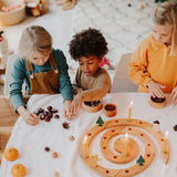 Three children sitting around a Grimm's natural spiral celebration ring with candles, acorns, pinecones, and small trees in a bright indoor room.