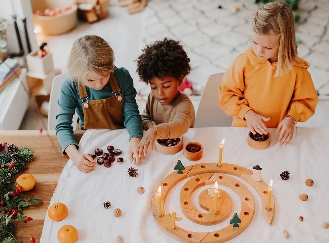 Three children sitting around a Grimm's natural spiral celebration ring with candles, acorns, pinecones, and small trees in a bright indoor room.