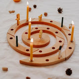 A Grimm's spiral wooden celebration ring with multiple lit yellow candles, styled on a white fabric surface with scattered pinecones and small pumpkins in the background.