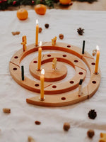 A Grimm's spiral wooden celebration ring with multiple lit yellow candles, styled on a white fabric surface with scattered pinecones and small pumpkins in the background.