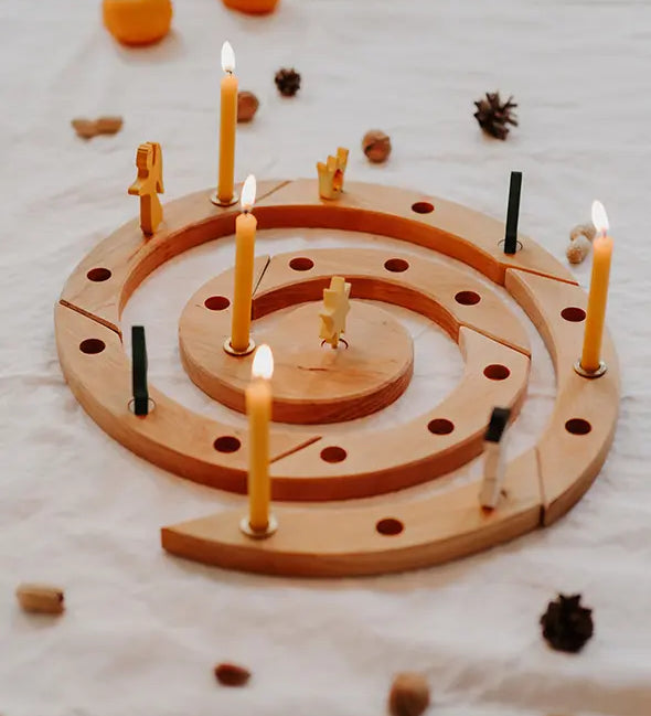 A Grimm's spiral wooden celebration ring with multiple lit yellow candles, styled on a white fabric surface with scattered pinecones and small pumpkins in the background.