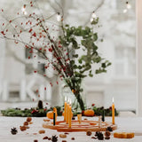 An autumnal tabletop arrangement with a Grimm's natural spiral celebration ring holding several lit yellow taper candles, surrounded by pinecones, acorns, small pumpkins, and a glass vase with greenery and red berries in front of a softly blurred window.